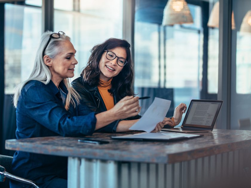 Duas mulheres em reunião de negócios analisando documentos e laptop.